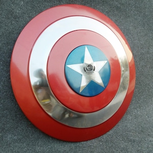 Op de afbeelding: Een rood, wit en blauw Captain America schild met een zilveren metalen rand. Het schild heeft een vijfpuntige ster in het midden met een blauwe achtergrond. De ster heeft een klein logo in het midden.