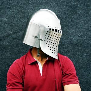 Op de afbeelding: Een zilverkleurige middeleeuwse helm met een vizier met talrijke kleine, gelijkmatig verdeelde gaten. De helm wordt gedragen over een rood poloshirt met een witte kraag. De achtergrond is een donkergrijze, getextureerde ondergrond.