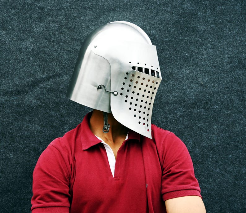 Op de afbeelding: Een zilverkleurige middeleeuwse helm met een vizier met talrijke kleine, gelijkmatig verdeelde gaten. De helm wordt gedragen over een rood poloshirt met een witte kraag. De achtergrond is een donkergrijze, getextureerde ondergrond.