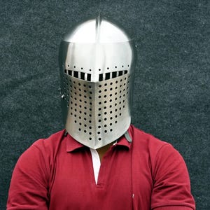 Op de afbeelding: Een gepolijste, zilverkleurige middeleeuwse helm met een volledig gezichtsvizier. Het vizier is geperforeerd met talrijke kleine gaatjes voor zicht en ventilatie. De helm wordt gedragen over een rood poloshirt met een witte kraag.