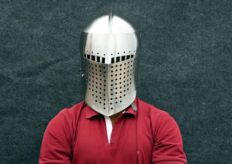 Op de afbeelding: Een gepolijste, zilverkleurige middeleeuwse helm met een volledig gezichtsvizier. Het vizier is geperforeerd met talrijke kleine gaatjes voor zicht en ventilatie. De helm wordt gedragen over een rood poloshirt met een witte kraag.
