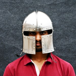 May include: A polished silver metal helmet with a rounded crown and rectangular face protection. The helmet features rivets and a small spike. The person is wearing a red polo shirt.