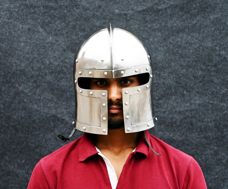 May include: A polished silver metal helmet with a rounded crown and rectangular face protection. The helmet features rivets and a small spike. The person is wearing a red polo shirt.