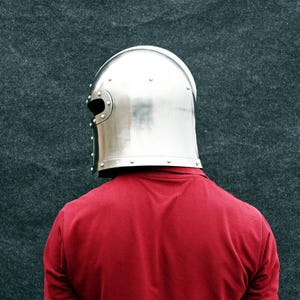 May include: A polished, silver metal helmet with a rounded crown and eye slits. The helmet is worn over a red top. The background is a dark grey, textured surface. The helmet is a medieval style.