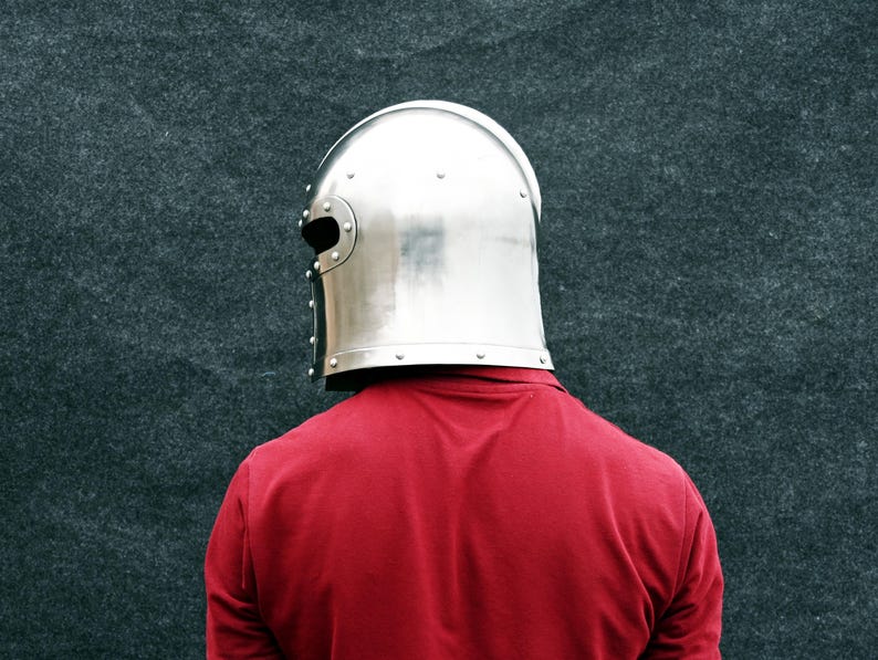 May include: A polished, silver metal helmet with a rounded crown and eye slits. The helmet is worn over a red top. The background is a dark grey, textured surface. The helmet is a medieval style.