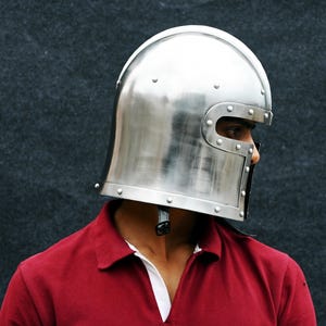 May include: A shiny silver medieval helmet with riveted detailing. The helmet has a curved top and a face guard with eye slits. The person is wearing a red polo shirt with a white collar.