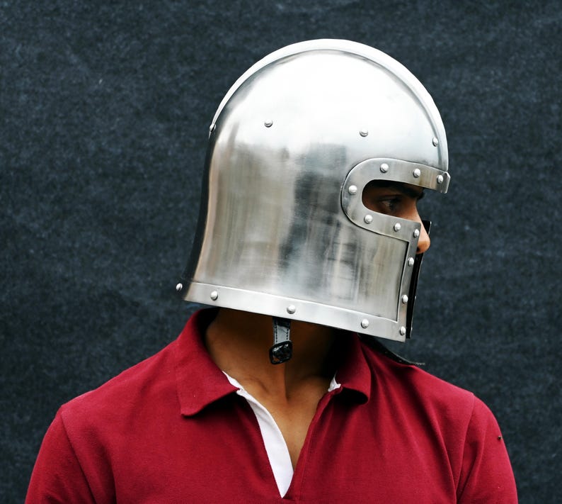 May include: A shiny silver medieval helmet with riveted detailing. The helmet has a curved top and a face guard with eye slits. The person is wearing a red polo shirt with a white collar.