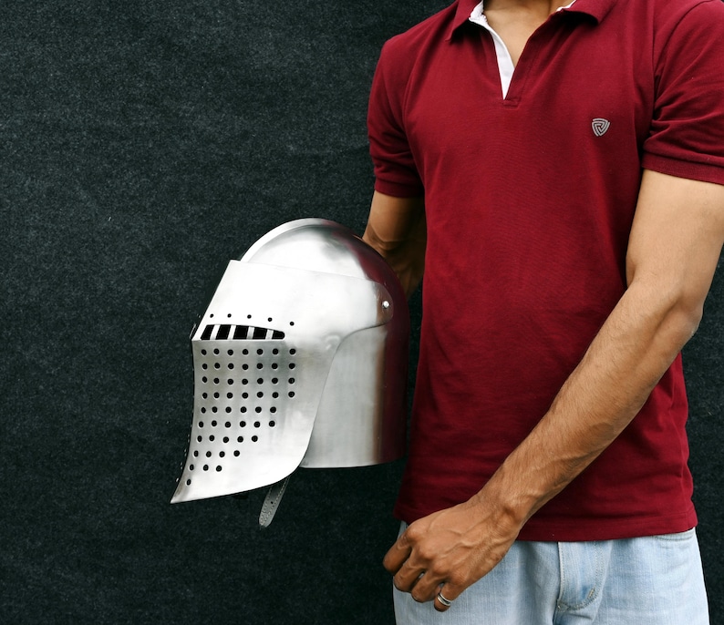 Op de afbeelding: Een gepolijste zilveren ridderhelm met een vizier met kleine gaatjes. De helm wordt tegen een donkere achtergrond gehouden. Het ontwerp van de helm doet denken aan middeleeuwse harnassen, geschikt voor historische re-enactments of kostuumevenementen.