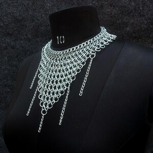 以下が含まれることがあります： 黒いマネキンに展示されたシルバーのチェーンメイルネックレス。 ネックレスは首の周りに織り込まれたデザインで、三角形の形を形成するぶら下がっているチェーンが付いています。 マネキンは首に番号「10」が付いています。