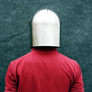 Op de afbeelding: Een glanzende, zilveren metalen helm met een afgeronde bovenkant en een verticale richel. De helm wordt gedragen over een rood shirt met lange mouwen. De achtergrond is een getextureerd, donkergroen oppervlak.