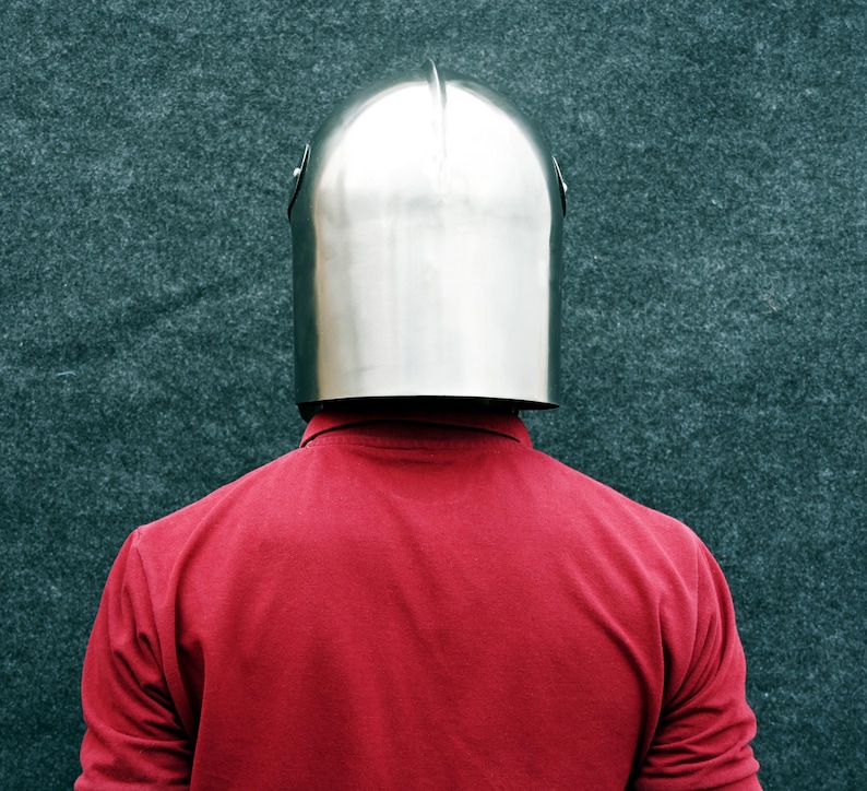 Op de afbeelding: Een glanzende, zilveren metalen helm met een afgeronde bovenkant en een verticale richel. De helm wordt gedragen over een rood shirt met lange mouwen. De achtergrond is een getextureerd, donkergroen oppervlak.