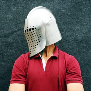 Op de afbeelding: Een zilverkleurige middeleeuwse helm met een vizier met talrijke kleine gaatjes. De helm wordt gedragen over een rood poloshirt met een witte kraag. De achtergrond is een donkergrijze, getextureerde ondergrond.