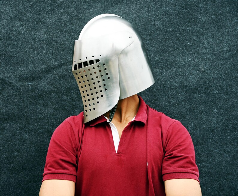 Op de afbeelding: Een zilverkleurige middeleeuwse helm met een vizier met talrijke kleine gaatjes. De helm wordt gedragen over een rood poloshirt met een witte kraag. De achtergrond is een donkergrijze, getextureerde ondergrond.