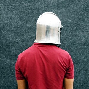 May include: A polished, silver metal helmet with rivets is worn over a red polo shirt. The helmet is a medieval style, covering the head and neck. The background is a textured, dark grey surface.
