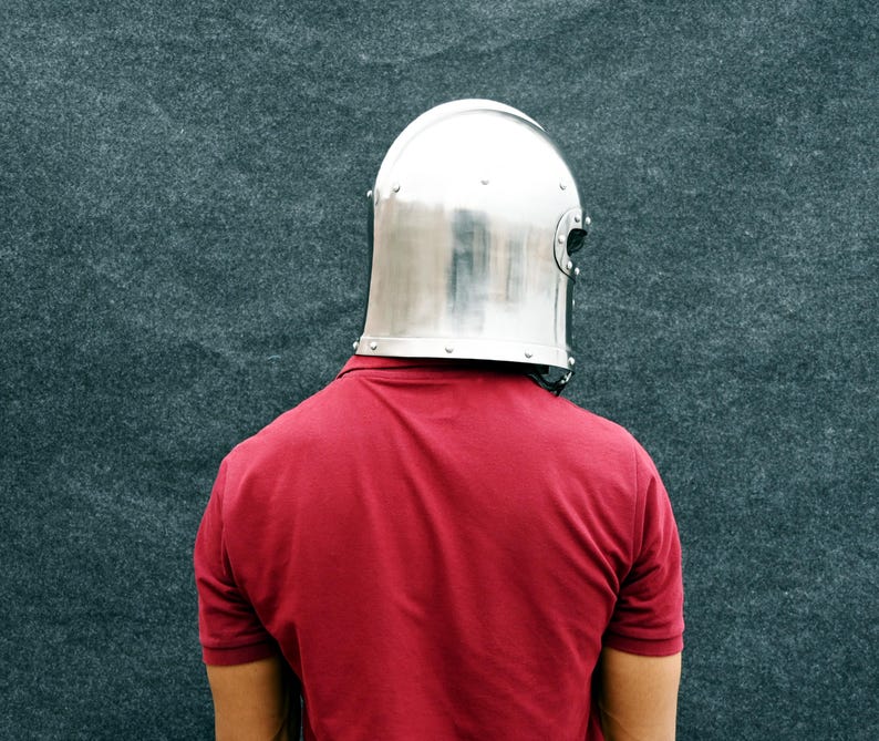 May include: A polished, silver metal helmet with rivets is worn over a red polo shirt. The helmet is a medieval style, covering the head and neck. The background is a textured, dark grey surface.