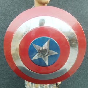 Op de afbeelding: Een rood, wit en blauw Captain America schild met een zilveren metalen rand. Het schild heeft een vijfpuntige ster in het midden met een logo dat "Coastal Designs" luidt.