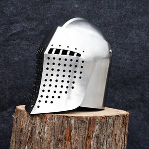 Op de afbeelding: Een glanzende zilveren ridderhelm met een geperforeerd vizier en horizontale oogspleten. De helm staat op een verweerde houten stronk tegen een donkergrijze achtergrond. Het ontwerp van de helm doet denken aan middeleeuwse harnassen.