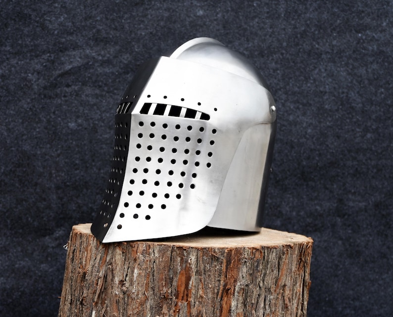 Op de afbeelding: Een glanzende zilveren ridderhelm met een geperforeerd vizier en horizontale oogspleten. De helm staat op een verweerde houten stronk tegen een donkergrijze achtergrond. Het ontwerp van de helm doet denken aan middeleeuwse harnassen.