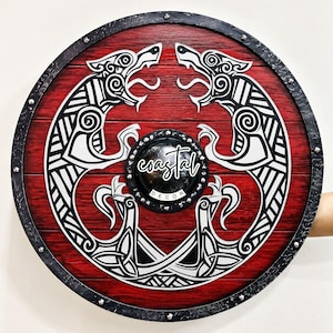 May include: A circular wooden shield featuring a deep red stained finish with a black and white Norse-style wolf design. The centre has a black metal boss with the text coastal DECOR. The rim is black with silver-toned metal rivets.