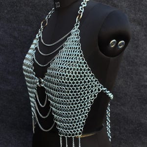 May include: A silver chainmail top with a halter neck design. The top is constructed of small, interlocking metal rings, with multiple strands of chains hanging from the neckline and the bottom edge. The garment is displayed on a black mannequin.
