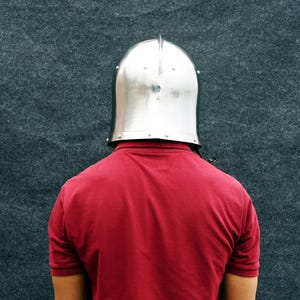 May include: A polished silver metal helmet with a central ridge, worn over a red polo shirt. The background is a dark grey, textured surface.