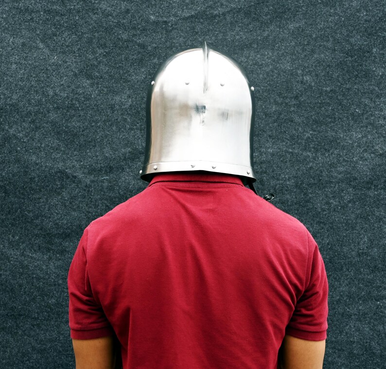 May include: A polished silver metal helmet with a central ridge, worn over a red polo shirt. The background is a dark grey, textured surface.