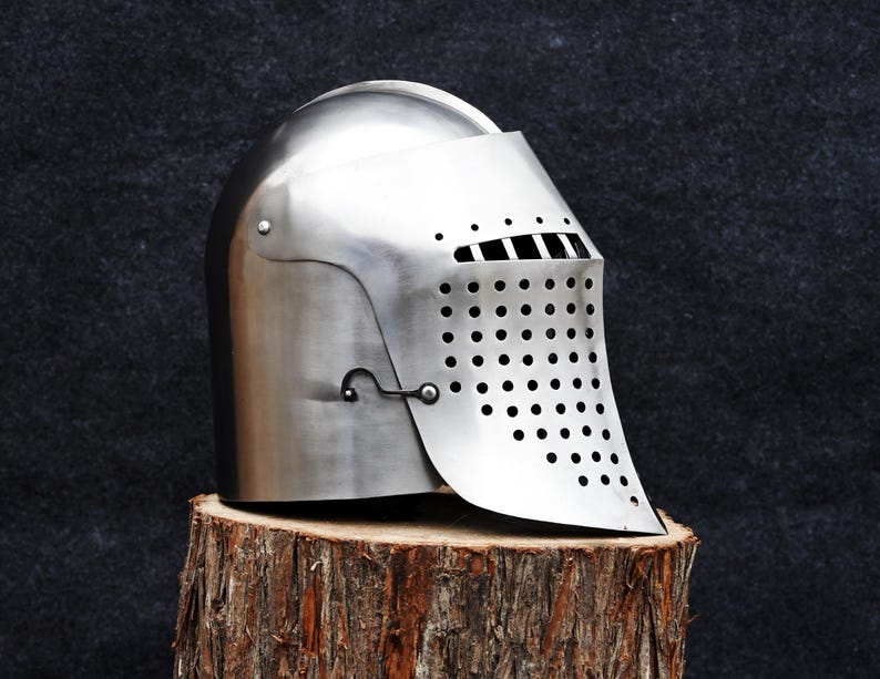 Op de afbeelding: Een gepolijste zilveren middeleeuwse helm met een vizier met talrijke kleine gaatjes. De helm staat op een rustieke houten stronk tegen een donkere achtergrond, wat het historische ontwerp benadrukt.
