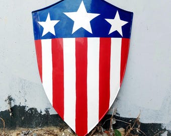Puede incluir: Un escudo con diseño patriótico, con una sección superior azul con tres estrellas blancas. La parte inferior tiene rayas verticales rojas y blancas. Este escudo es probablemente un accesorio de disfraz o un artículo decorativo.