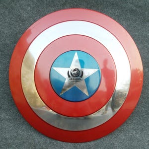 Op de afbeelding: Een rood, wit en blauw Captain America schild met een zilveren metalen rand en een vijfpuntige ster in het midden. De ster heeft een blauwe achtergrond en een zilveren metalen rand. Het schild heeft de tekst "Cosplay" in zwarte letters binnen de ster.