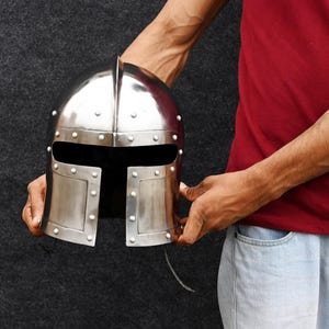 May include: A shiny silver medieval helmet with rivet detailing. The helmet has a central ridge and a face opening. The metal helmet is held against a dark background.