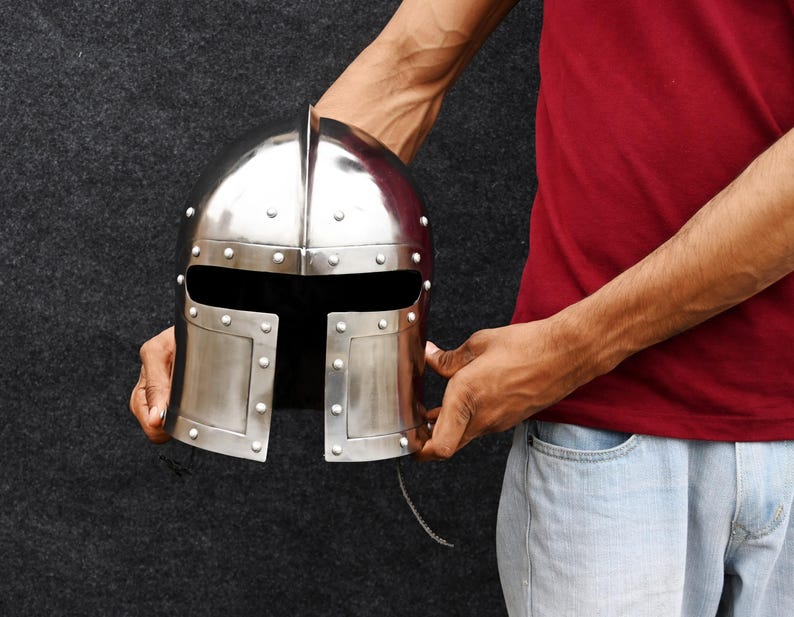 May include: A shiny silver medieval helmet with rivet detailing. The helmet has a central ridge and a face opening. The metal helmet is held against a dark background.