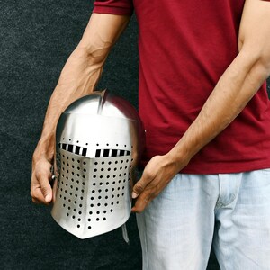 Op de afbeelding: Een zilverkleurige middeleeuwse helm met een vizier met rechthoekige en cirkelvormige perforaties. De helm wordt vastgehouden door een persoon die een rood shirt en lichtblauwe jeans draagt.