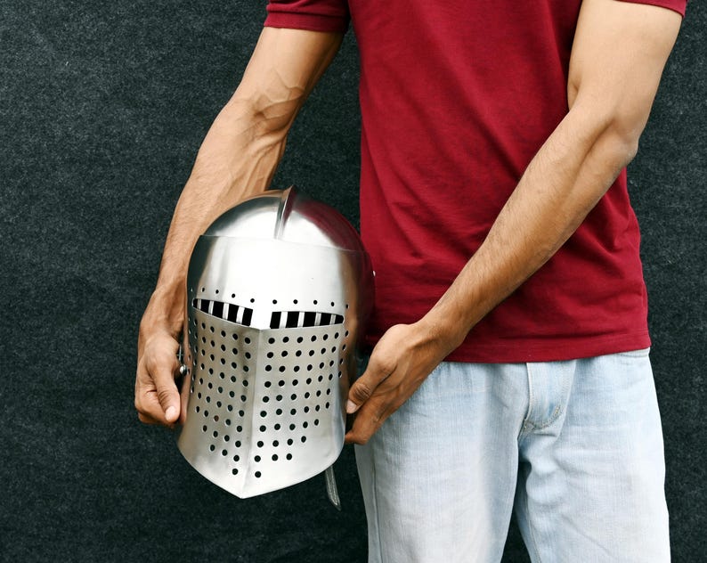 Op de afbeelding: Een zilverkleurige middeleeuwse helm met een vizier met rechthoekige en cirkelvormige perforaties. De helm wordt vastgehouden door een persoon die een rood shirt en lichtblauwe jeans draagt.