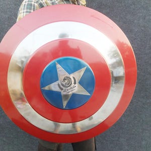Op de afbeelding: Een rood, wit en blauw Captain America schild met een zilveren metalen rand. Het schild heeft een vijfpuntige ster in het midden met een drakenlogo en de tekst "Coastal", "Decor" en "com".
