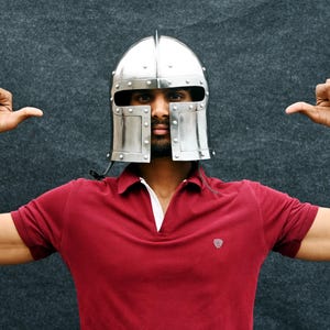 May include: A silver metal helmet with rivets, worn over a red polo shirt. The person is making a thumbs-up gesture with both hands. The background is a dark grey.