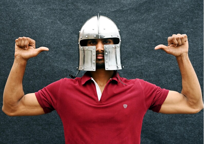 May include: A silver metal helmet with rivets, worn over a red polo shirt. The person is making a thumbs-up gesture with both hands. The background is a dark grey.