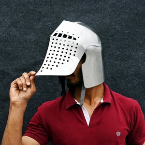 Op de afbeelding: Een zilverkleurige middeleeuwse helm met een vizier met talrijke kleine, gelijkmatig verdeelde gaten. De helm wordt omhooggehouden, waardoor het gezicht van de persoon zichtbaar wordt. De persoon draagt een rode polo.