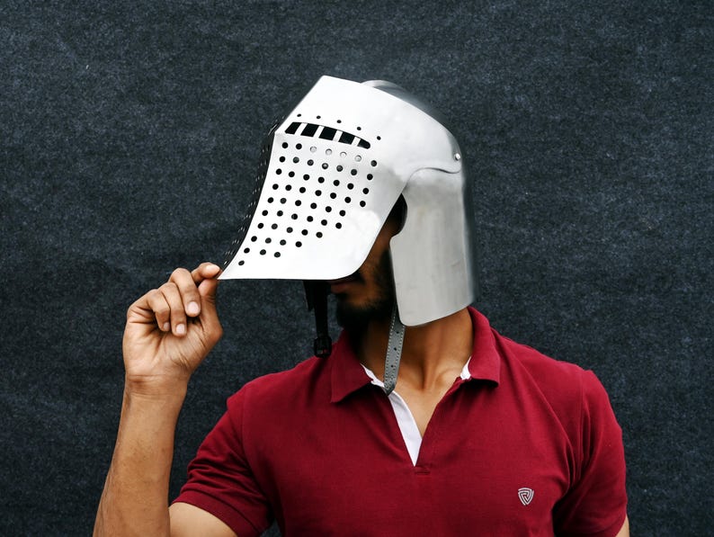 Op de afbeelding: Een zilverkleurige middeleeuwse helm met een vizier met talrijke kleine, gelijkmatig verdeelde gaten. De helm wordt omhooggehouden, waardoor het gezicht van de persoon zichtbaar wordt. De persoon draagt een rode polo.