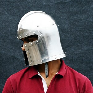 May include: A shiny silver medieval helmet with rivet detailing. The helmet has a rounded crown and a face guard with eye slits. The person is wearing a red polo shirt with a white collar.