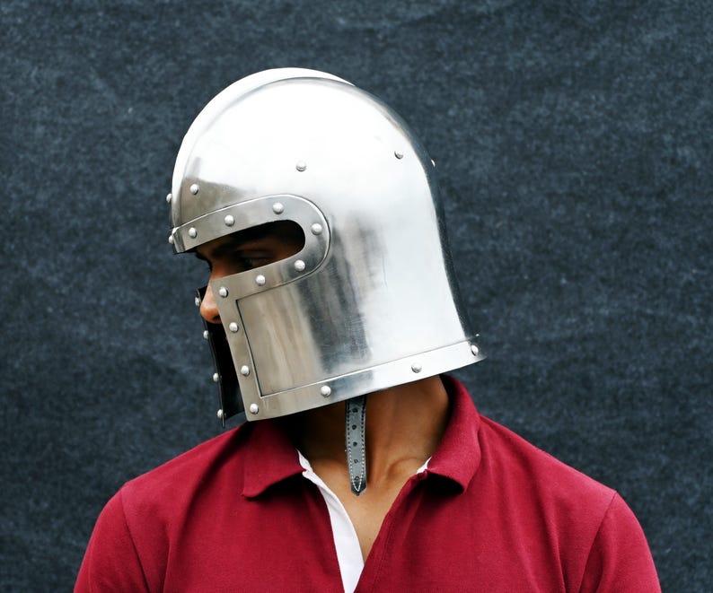 May include: A shiny silver medieval helmet with rivet detailing. The helmet has a rounded crown and a face guard with eye slits. The person is wearing a red polo shirt with a white collar.