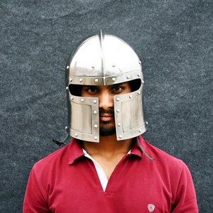 May include: A polished silver medieval helmet with a pointed top and riveted construction. The helmet features a face guard with rectangular openings for the eyes and a hinged lower section. The person is wearing a red polo shirt.