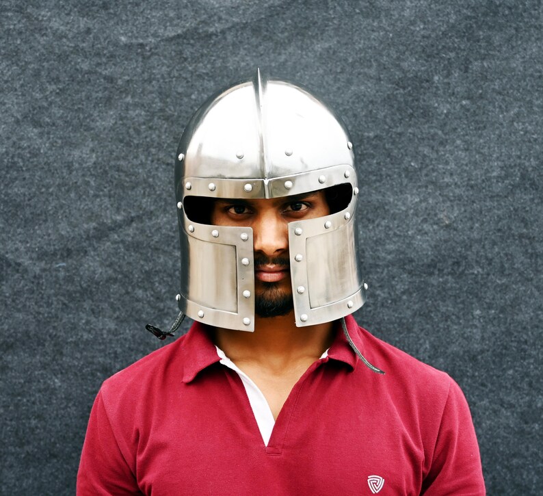 May include: A polished silver medieval helmet with a pointed top and riveted construction. The helmet features a face guard with rectangular openings for the eyes and a hinged lower section. The person is wearing a red polo shirt.