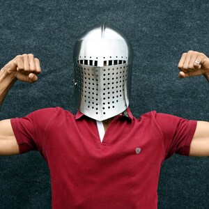 Op de afbeelding: Een glanzende, zilveren middeleeuwse helm met een geperforeerd vizier. De persoon draagt een rood poloshirt en buigt zijn armen, waarbij hij zijn biceps laat zien. De achtergrond is een donker, getextureerd oppervlak.