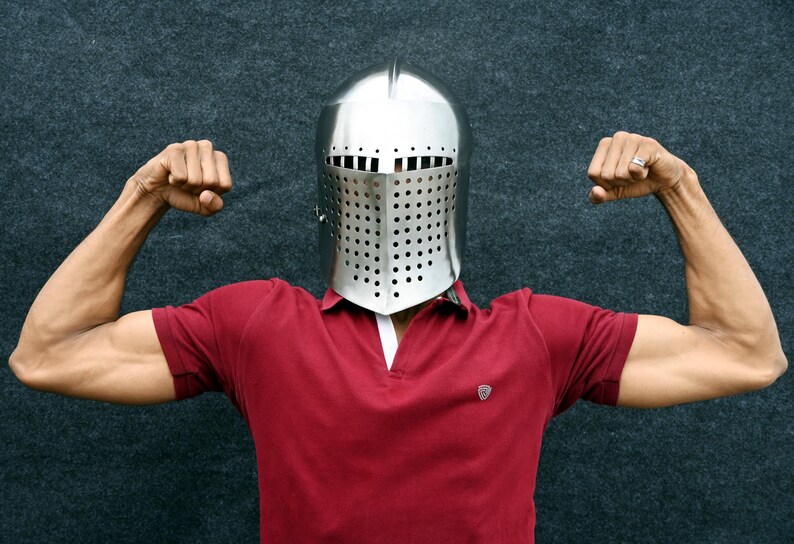 Op de afbeelding: Een glanzende, zilveren middeleeuwse helm met een geperforeerd vizier. De persoon draagt een rood poloshirt en buigt zijn armen, waarbij hij zijn biceps laat zien. De achtergrond is een donker, getextureerd oppervlak.