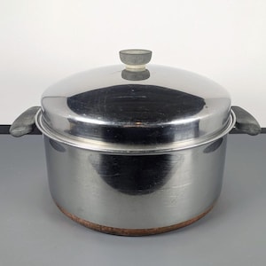 Peut inclure: Une marmite en acier inoxydable avec un couvercle assorti, dotée de deux poignées latérales gris foncé et d'un bouton gris assorti sur le couvercle. La base de la marmite présente une bande cuivrée et les surfaces métalliques sont brillantes.