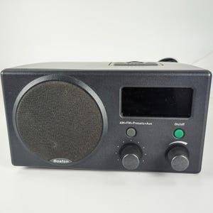 May include: A black Boston brand tabletop radio featuring a circular speaker grille on the left, a rectangular digital display screen, and a control panel with a grey button labelled AM>FM>Presets>Aux and a green button labelled On/off.