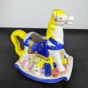 May include: A ceramic rocking horse figurine featuring a white body, bright yellow mane and tail, and a blue saddle with gold trim. The base is decorated with colourful wrapped gift boxes in shades of pink, green, and blue with gold accents.