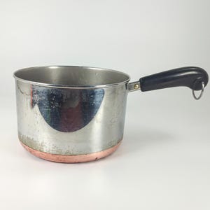 May include: A stainless steel saucepan with a copper-coloured base and a black handle. The handle features a small metal loop at the end for hanging. The exterior of the pot shows signs of wear and surface scratches on the polished metal finish.