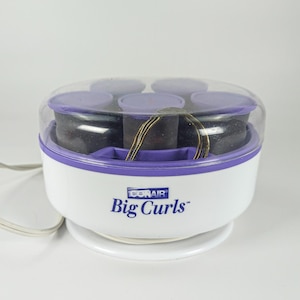 May include: A white and purple Conair Big Curls hair setter featuring a clear plastic lid, a circular base, and a white power cord. The unit contains several dark rollers and gold-toned metal clips arranged inside the heating compartment.