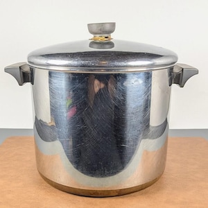 Peut inclure: Une grande marmite cylindrique en acier inoxydable avec une finition polie et réfléchissante présentant des rayures de surface. Elle possède deux poignées latérales gris foncé et un couvercle assorti avec un bouton central sur le dessus.